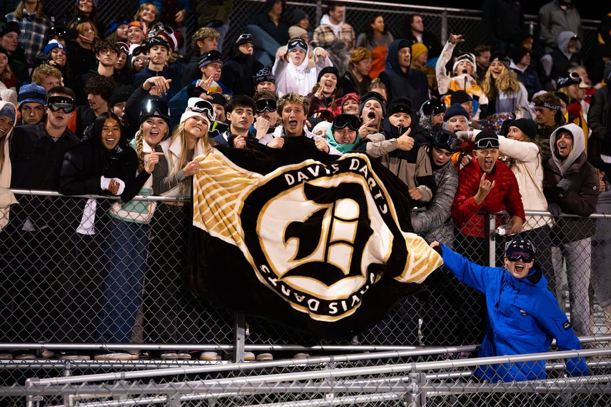 A packed high school student section holding up a custom Davis Darts mascot blanket at a cold-weather football game